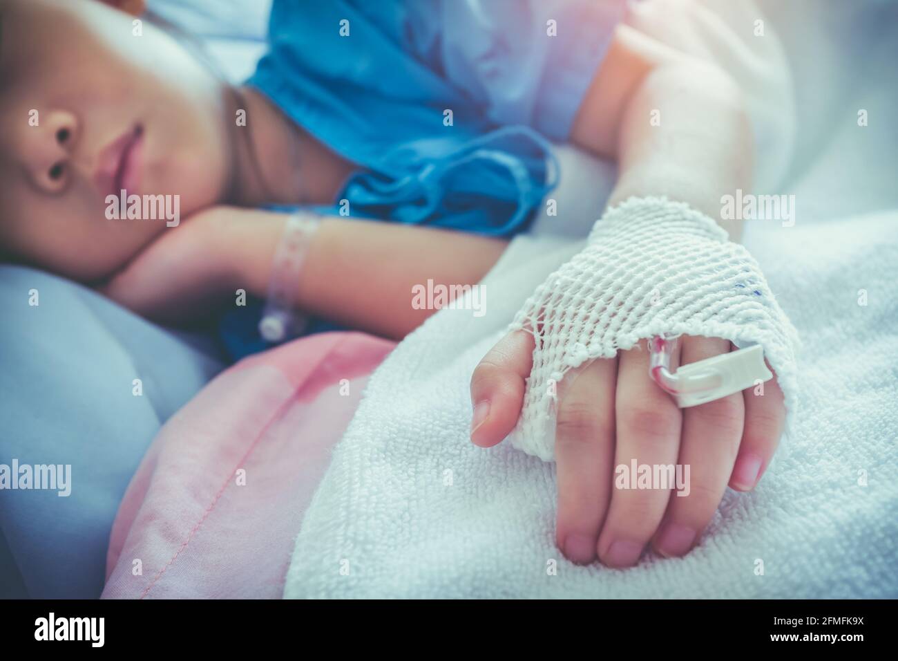 Illness asian child admitted in hospital with saline intravenous (IV) on hand. Girl sleeping at comfortable equipped hospital room with sunlight. Heal Stock Photo