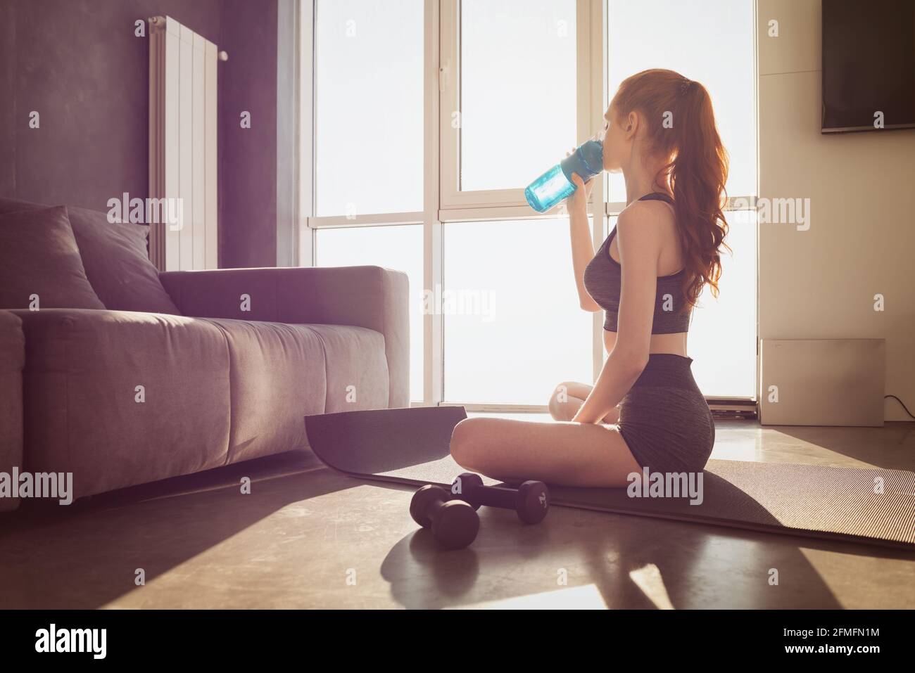 Young woman drinks water from the bottle after doing physical training exercises at home on the mat Stock Photo