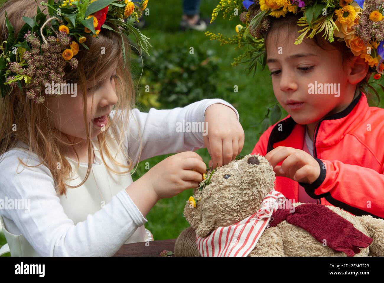 London, UK, 9 May 2021: School friends Emily, age 4 (cream dress) and Emelia, age 5 (orange cardigan), make a flower crowns to celebrate Garden Day, and one for the class teddy bear too. This event was at Chelsea Physic Garden and led by florist Fran Bailey of The Fresh Flower company. Anna Watson/Alamy Live News Stock Photo
