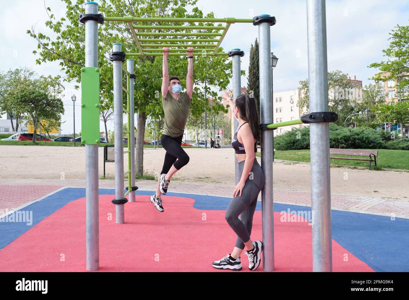 Two friends swinging in a monkey bar at a calisthenics park wearing protective face masks. Fitness outdoor. Stock Photo