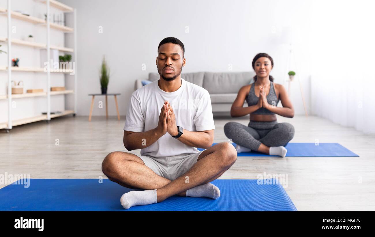 Black guy meditating with closed eyes, sitting in lotus pose, doing namaste gesture, practicing yoga with his girlfriend Stock Photo