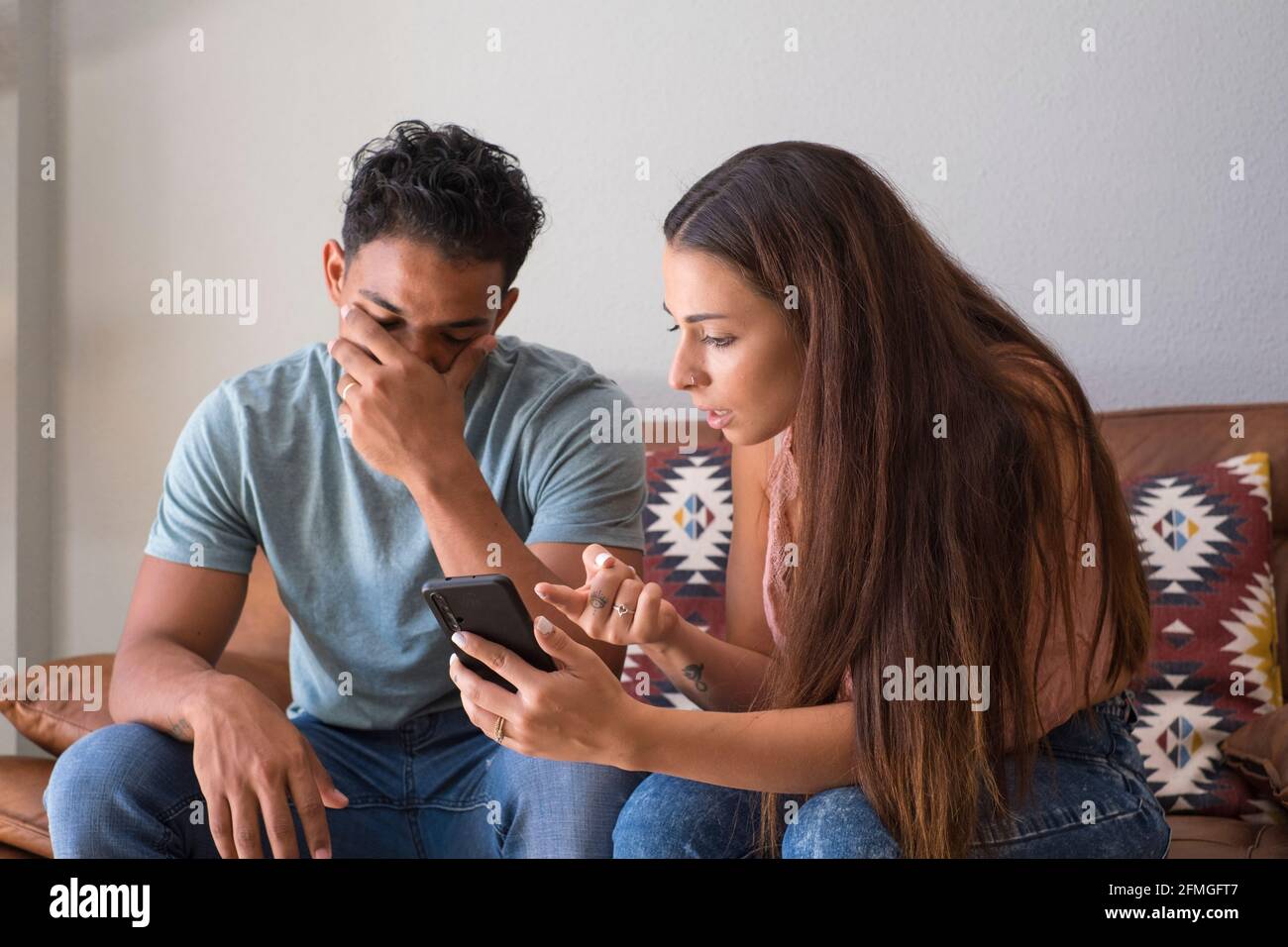 Infidelity. Jealous girlfriend Showing his Cheating boyfriend his Phone Demanding Explanation Sitting On Sofa Indoor at home. Wife caught her husband Stock Photo