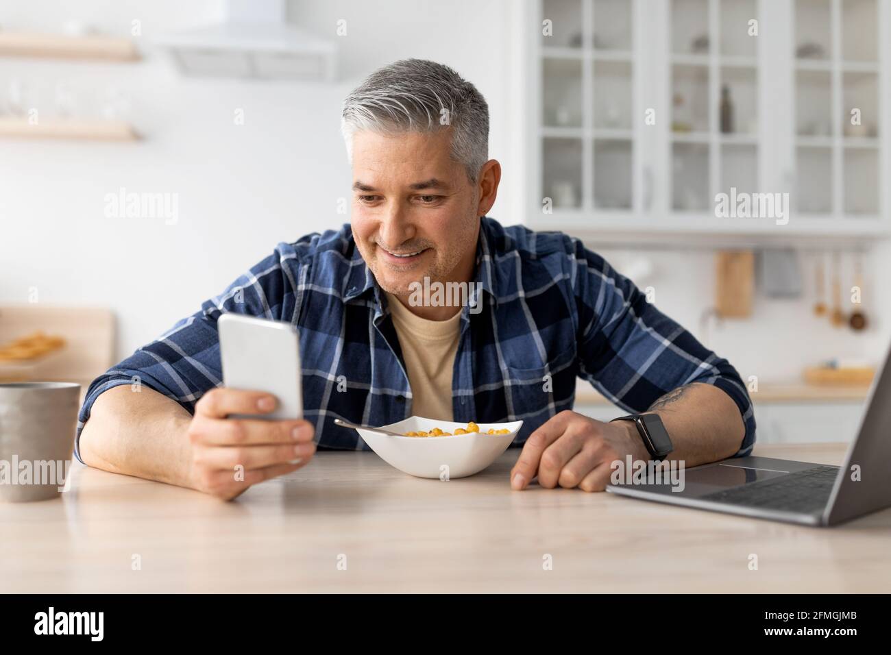 Matured man using smartphone while having healthy breakfast in kitchen at home, sitting at table and eating cereals Stock Photo