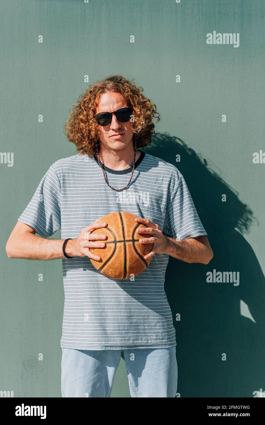 vertical portrait of a thin long-haired caucasian young man . He is holding a basketball ball with both hands. Looking at camera serious Stock Photo