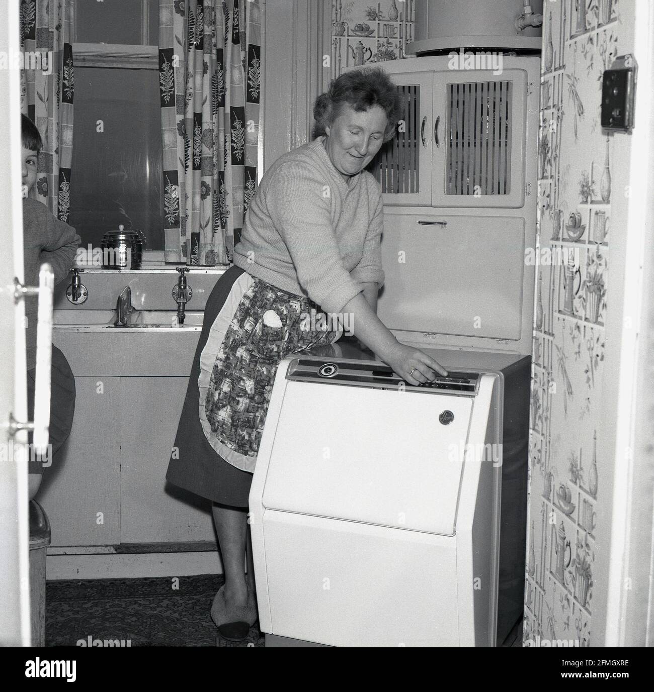 1960s, historical, inside a small kitchen, a blind or partially sighted lady, in an apron for doing her domestic duties, using a new 'braille' washing machine, Kelty, Fife, Scotland. The washing machine had controls in the tactile 'braille' writing system so it could be used by those who were visually impaired. Braille is a touch system in which raised dots or bumps represent the letters of the alphabet. Stock Photo