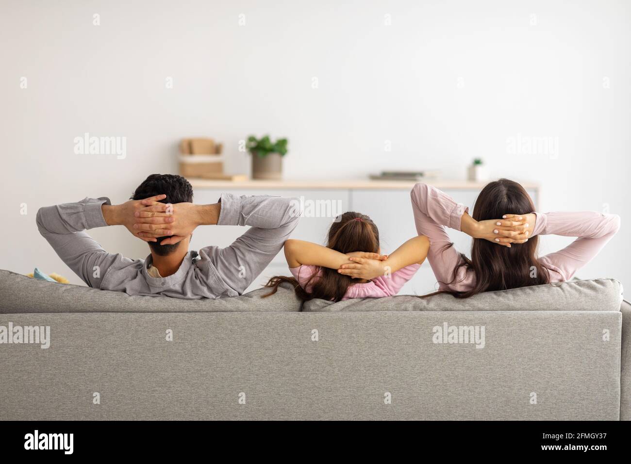 Leisure time. Back view of relaxed parents and child with arms behind head sitting on couch and resting at home Stock Photo