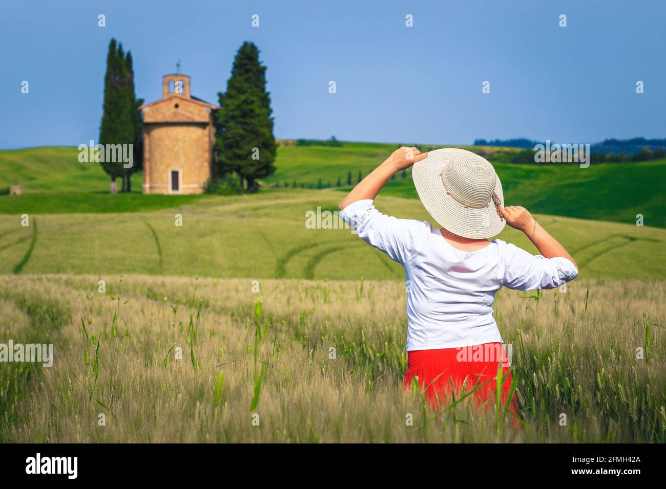 Beautiful cheerful woman in red skirt and straw hat enjoying the freedom and view. Countryside scenery with grain fields and Vitaleta chapel, Tuscany, Stock Photo
