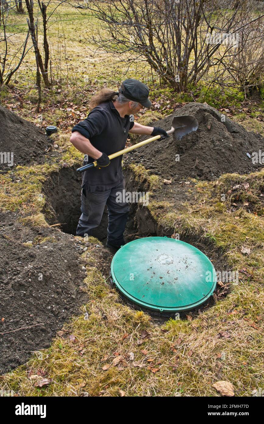 Man digging up home septic tank with a shovel with an access hatch in the foreground. Stock Photo