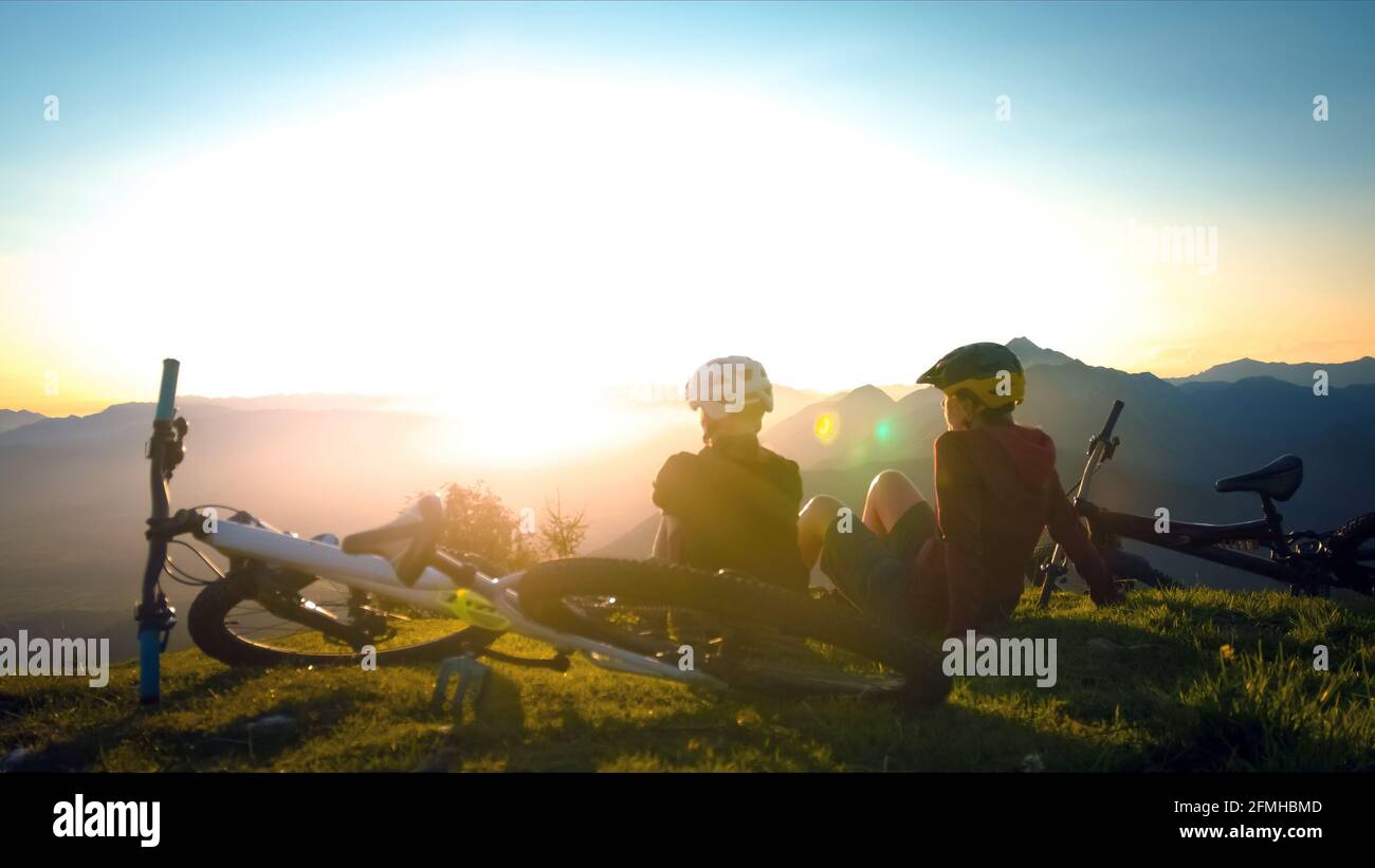 Girls sitting on mountain with bycicles looking at sunset and talking. Enjoying in beautiful nature. Stock Photo