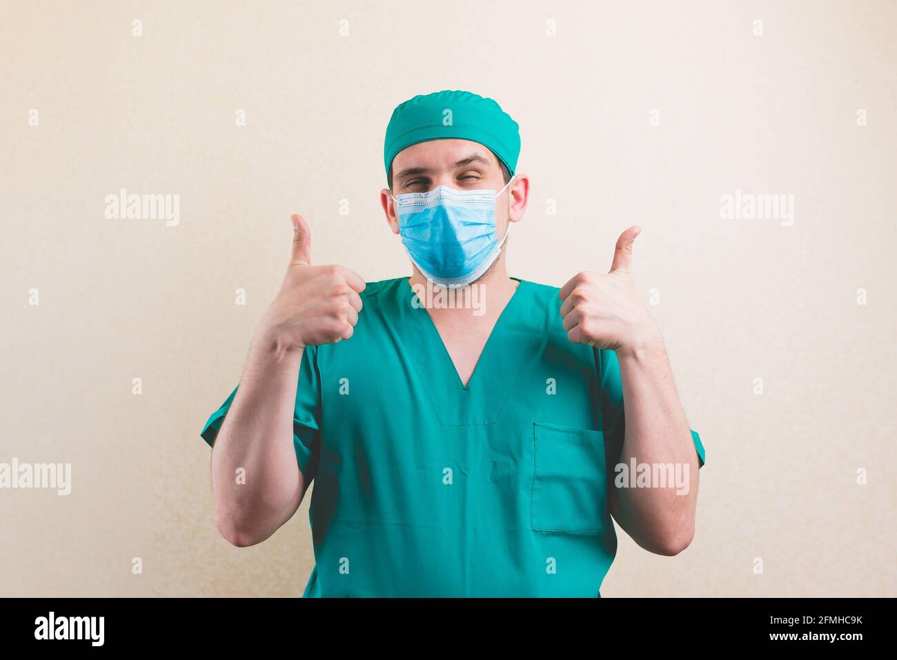 Young squinting doctor portrait with thumbs up in green surgeon suit and protective breathing mask on the yellow background isolated Stock Photo