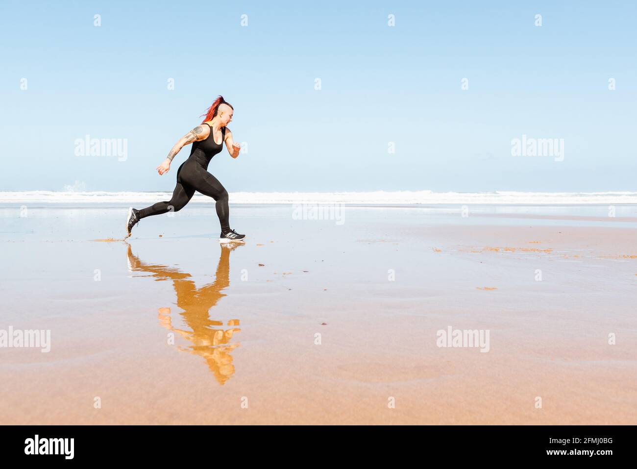 Side view of active female jogger with tattoos running on ocean shore ...