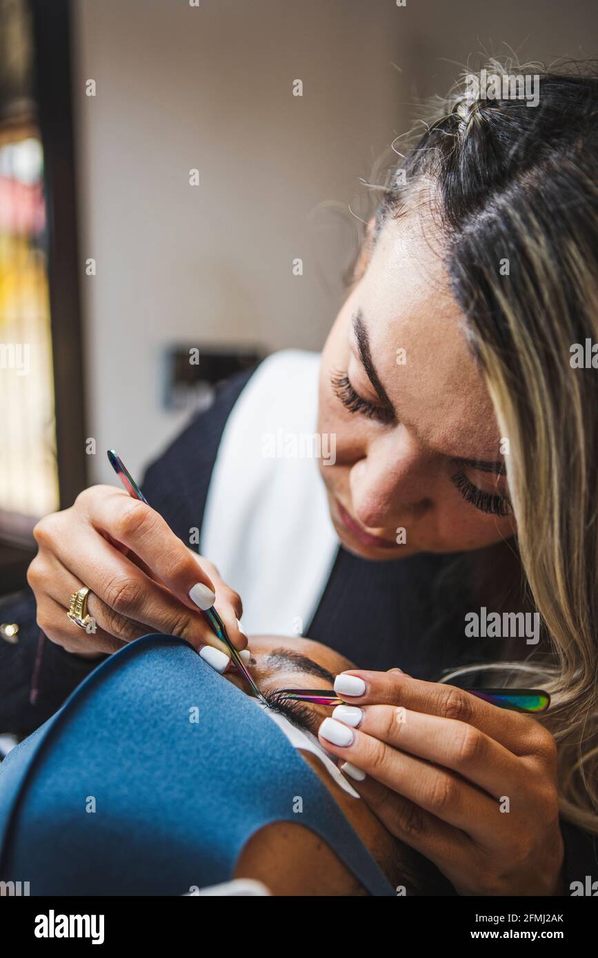 Cosmetologist with tweezers applying fake eyelashes for extension on eye of ethnic client with face protective mask in salon during coronavirus pandem Stock Photo