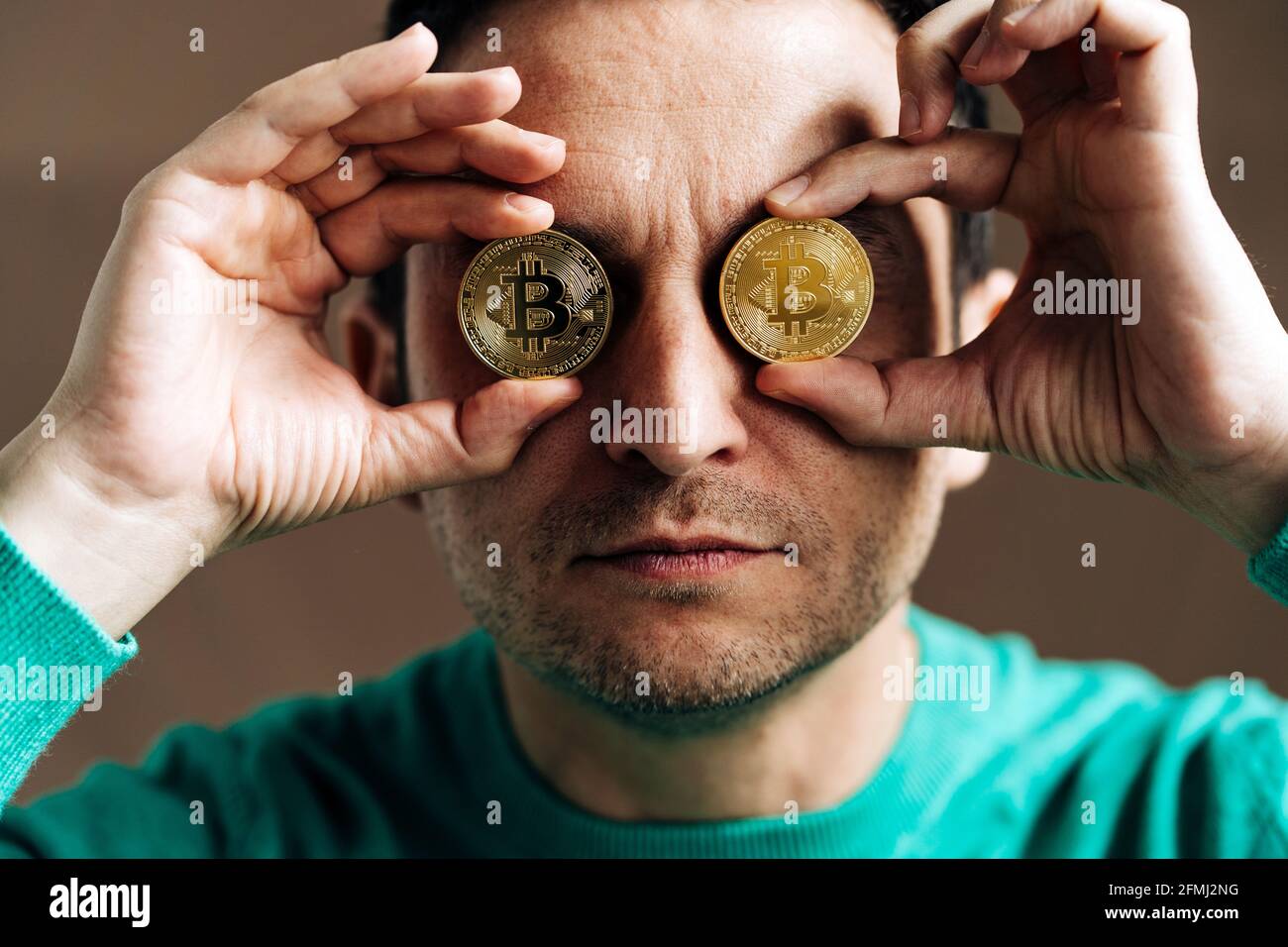 Male broker in green wear covering eyes with metal crypto coins on brown  background Stock Photo - Alamy