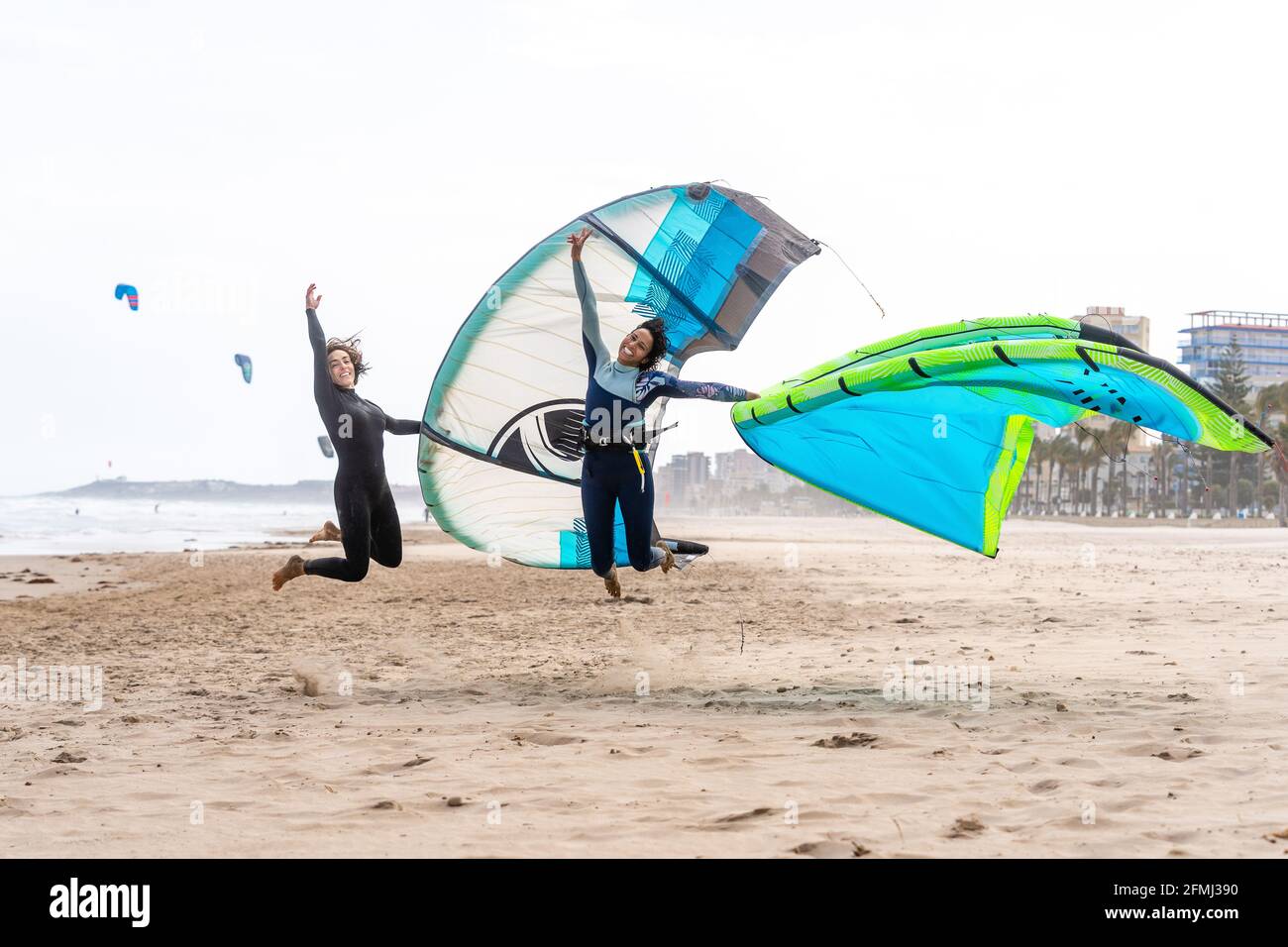 Cheerful multiethnic female kiters with power kites jumping above sandy ocean coast under white sky in city Stock Photo