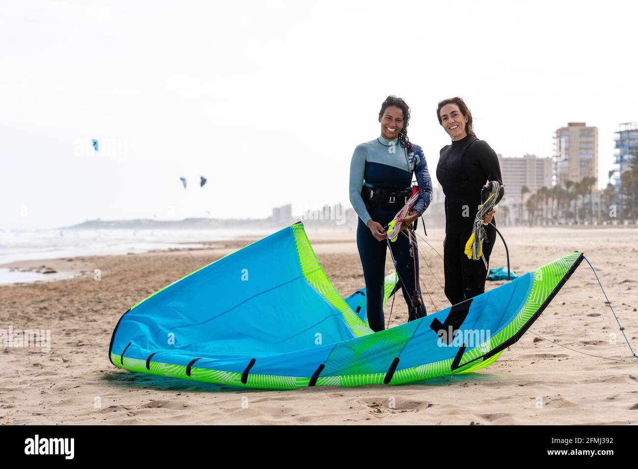 Cheerful multiethnic female kiters with power kites on sandy ocean coast under white sky in city Stock Photo