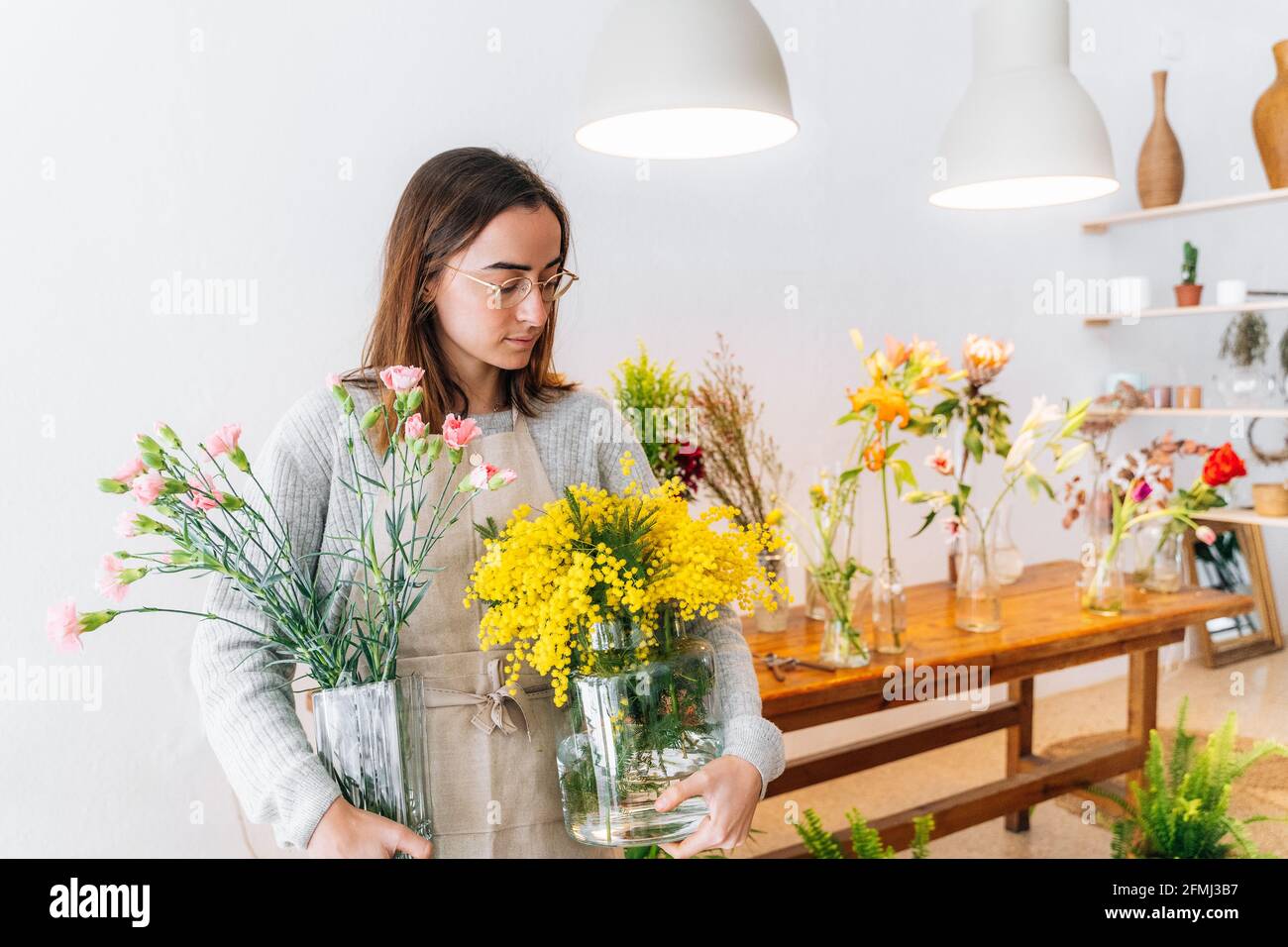 Calm young ethnic female florist in apron and eyeglasses holding glass vases with yellow mimosa and clove pink flowers in store Stock Photo