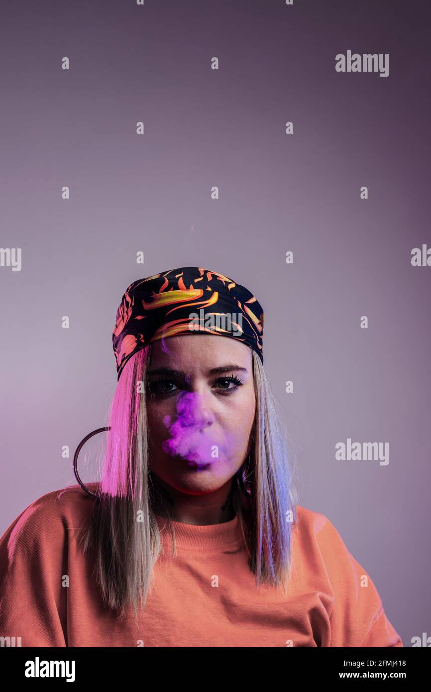Cool female in street style outfit smoking e cigarette and exhaling smoke through nose on purple background in studio with pink neon illumination Stock Photo