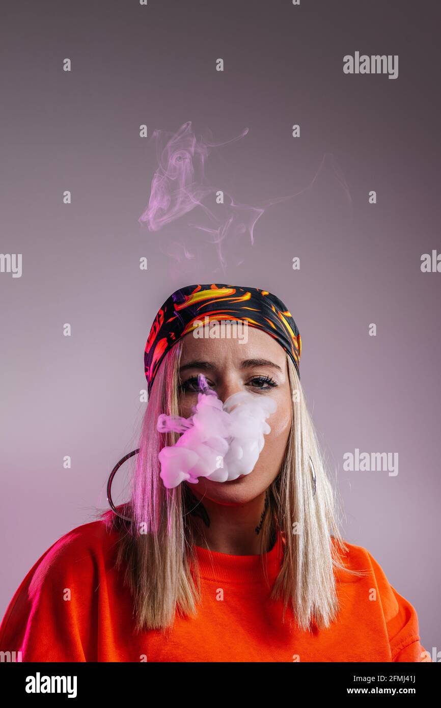 Cool female in street style outfit smoking e cigarette and exhaling smoke through nose on purple background in studio with pink neon illumination Stock Photo