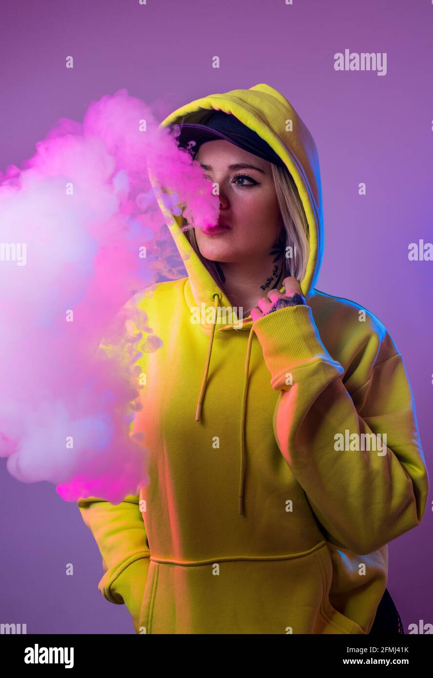 Confident female hipster in hoodie smoking e cigarette in studio on pink background and looking away Stock Photo