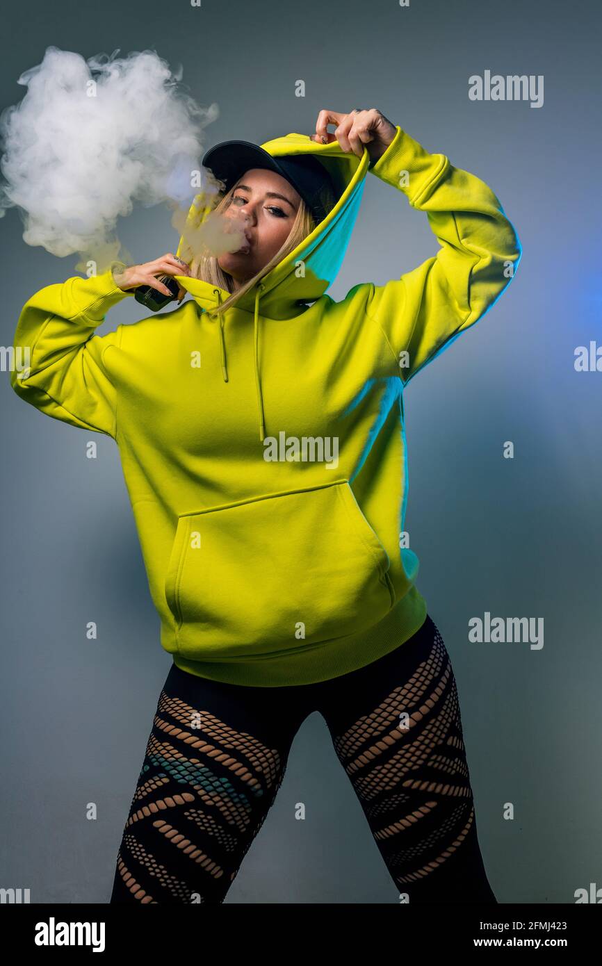 Confident female hipster in hoodie smoking e cigarette in studio on gray background and looking at camera Stock Photo