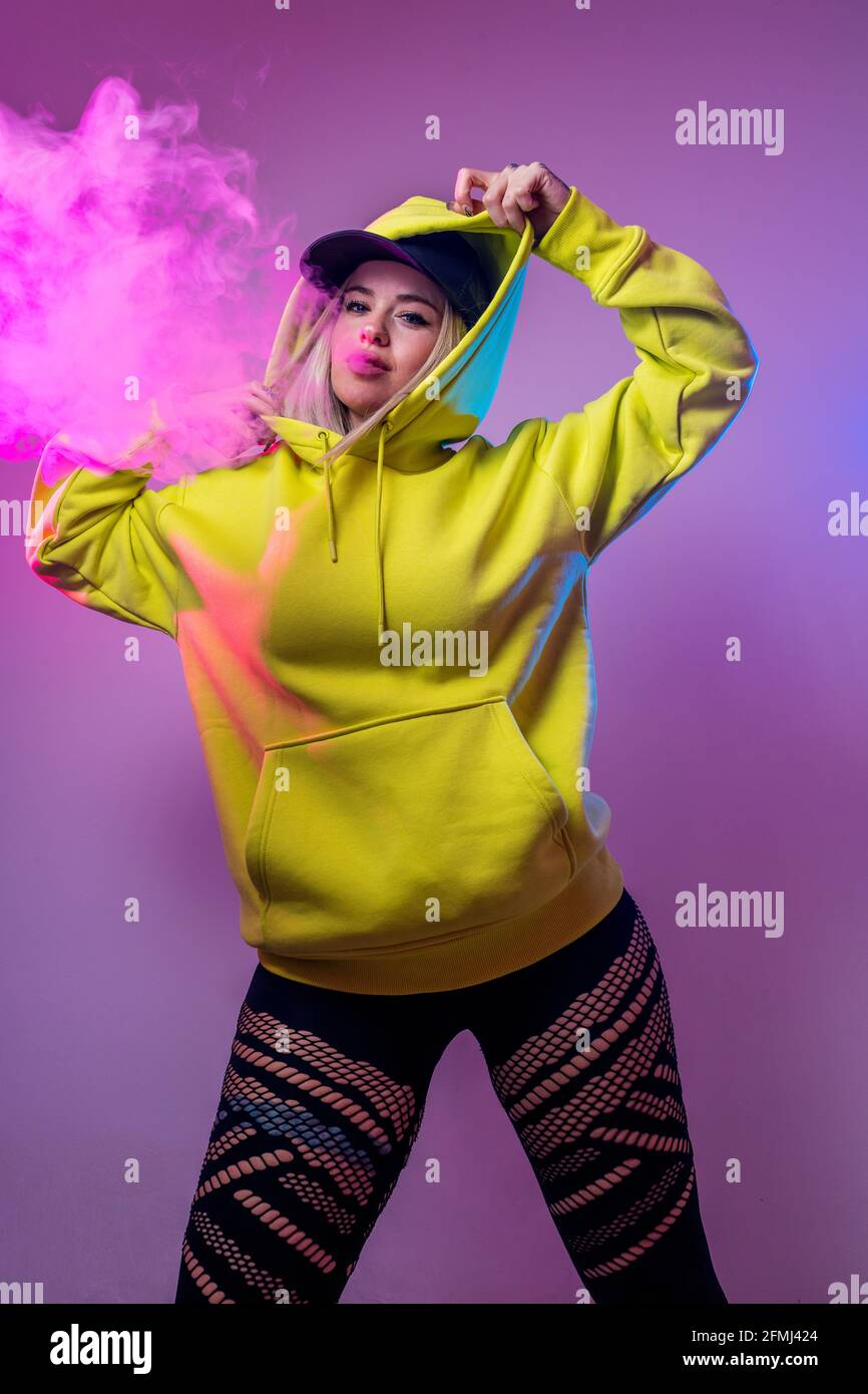 Confident female hipster in hoodie smoking e cigarette in studio on pink background and looking at camera Stock Photo