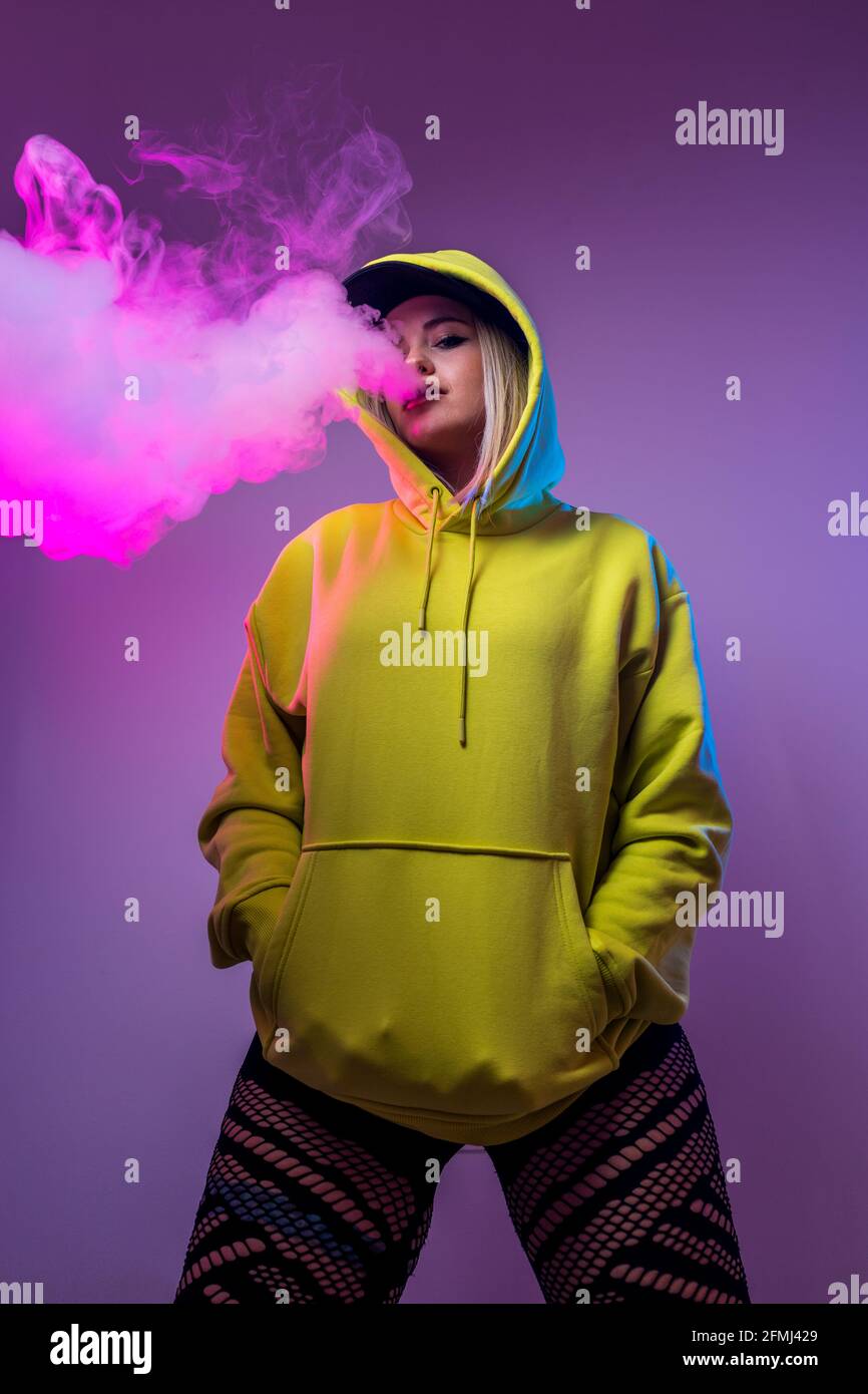 From below confident female hipster in hoodie smoking e cigarette in studio on pink background and looking at camera Stock Photo