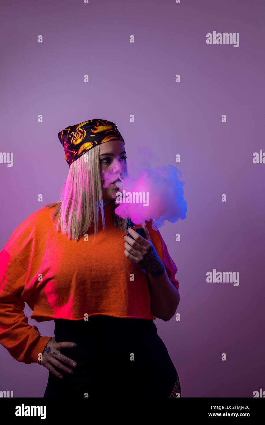Cool female in street style outfit smoking e cigarette and exhaling smoke through nose and mouth on purple background in studio with pink neon illumin Stock Photo