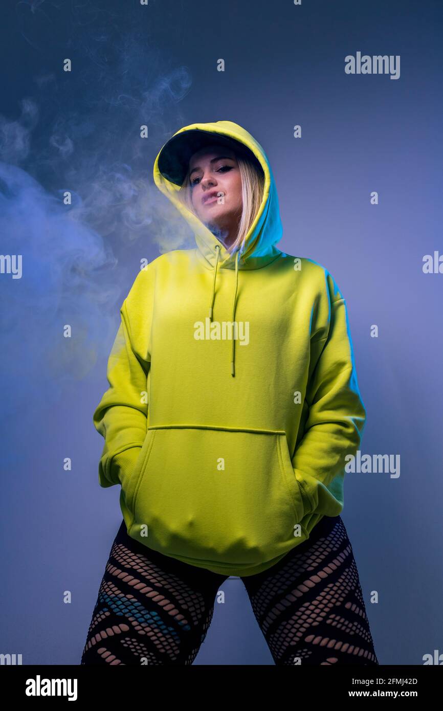 From below confident female hipster in hoodie smoking e cigarette in studio on gray background and looking at camera Stock Photo