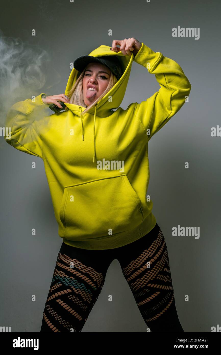 Confident female hipster in hoodie smoking vape in studio on gray background and looking at camera while sticking tongue out Stock Photo