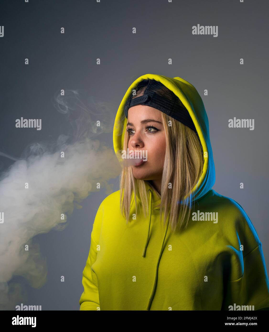 Confident female hipster in hoodie smoking e cigarette in studio on gray background and looking away Stock Photo