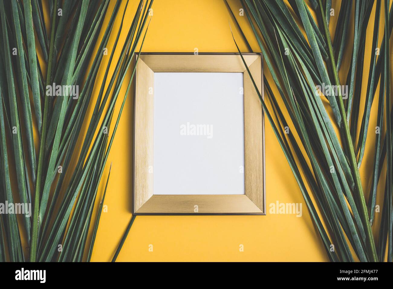 Wooden frame with white picture hanging on orange wall decorated with fresh green palm leaves at home Stock Photo