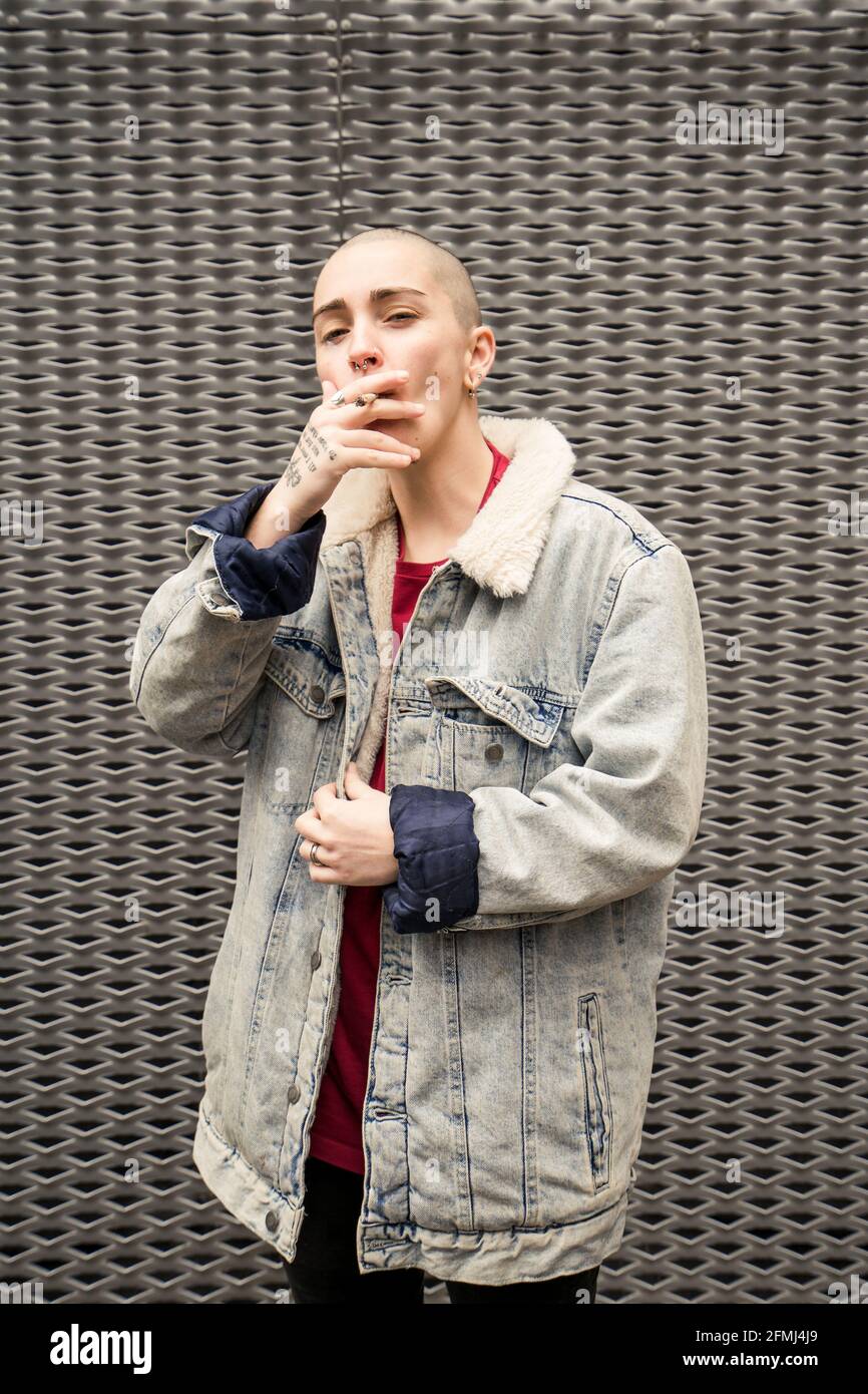 Self confident young transgender person in denim jacket smoking cigarette while looking at camera Stock Photo