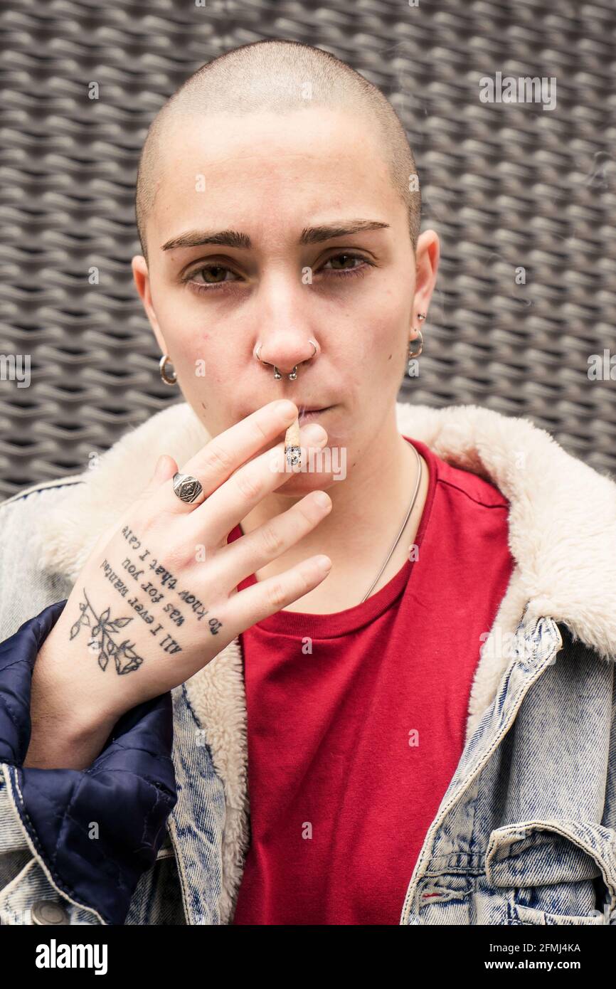 Self confident young transgender person in denim jacket smoking cigarette while looking at camera Stock Photo