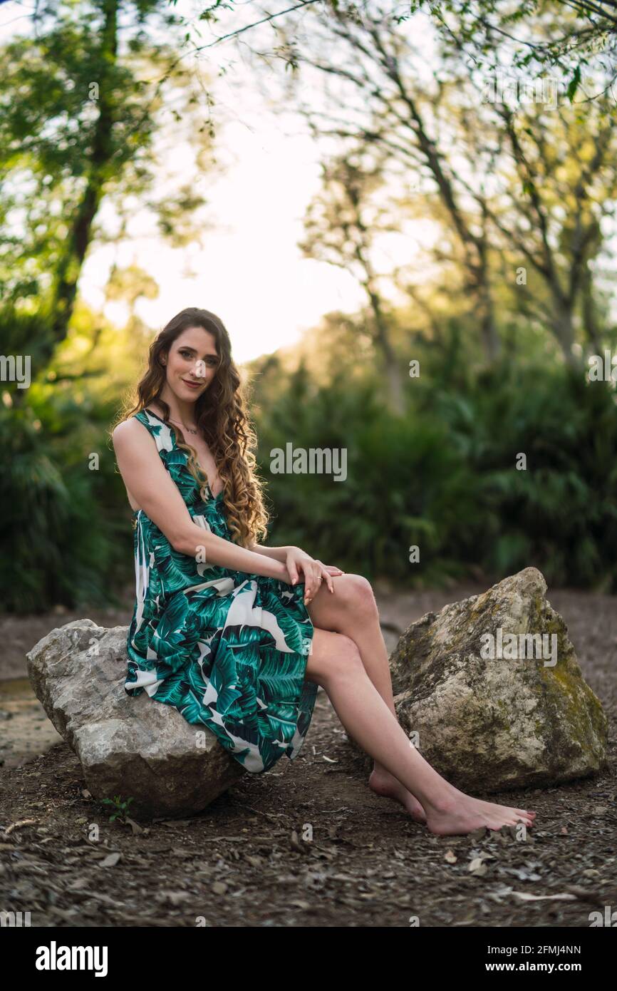 Full body content young female wearing trendy maxi sundress and looking at camera with smile while sitting on big stone in verdant sunny woodland Stock Photo