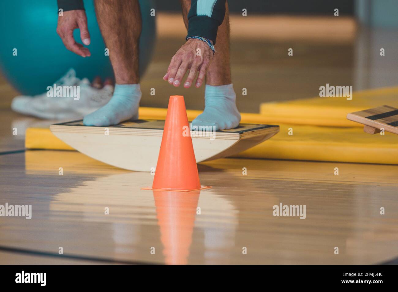 Male trainer seen Touching a cone while on wooden balance board. Different balance sports equipment is seen around in a gym indoors. Stock Photo
