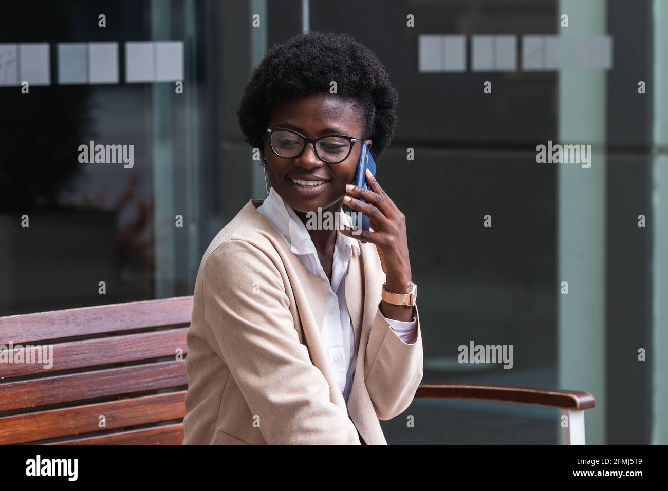 Young African American business lady with short hair wearing stylish suit  and glasses smiling during phone conversation while sitting on bench near  ur Stock Photo - Alamy