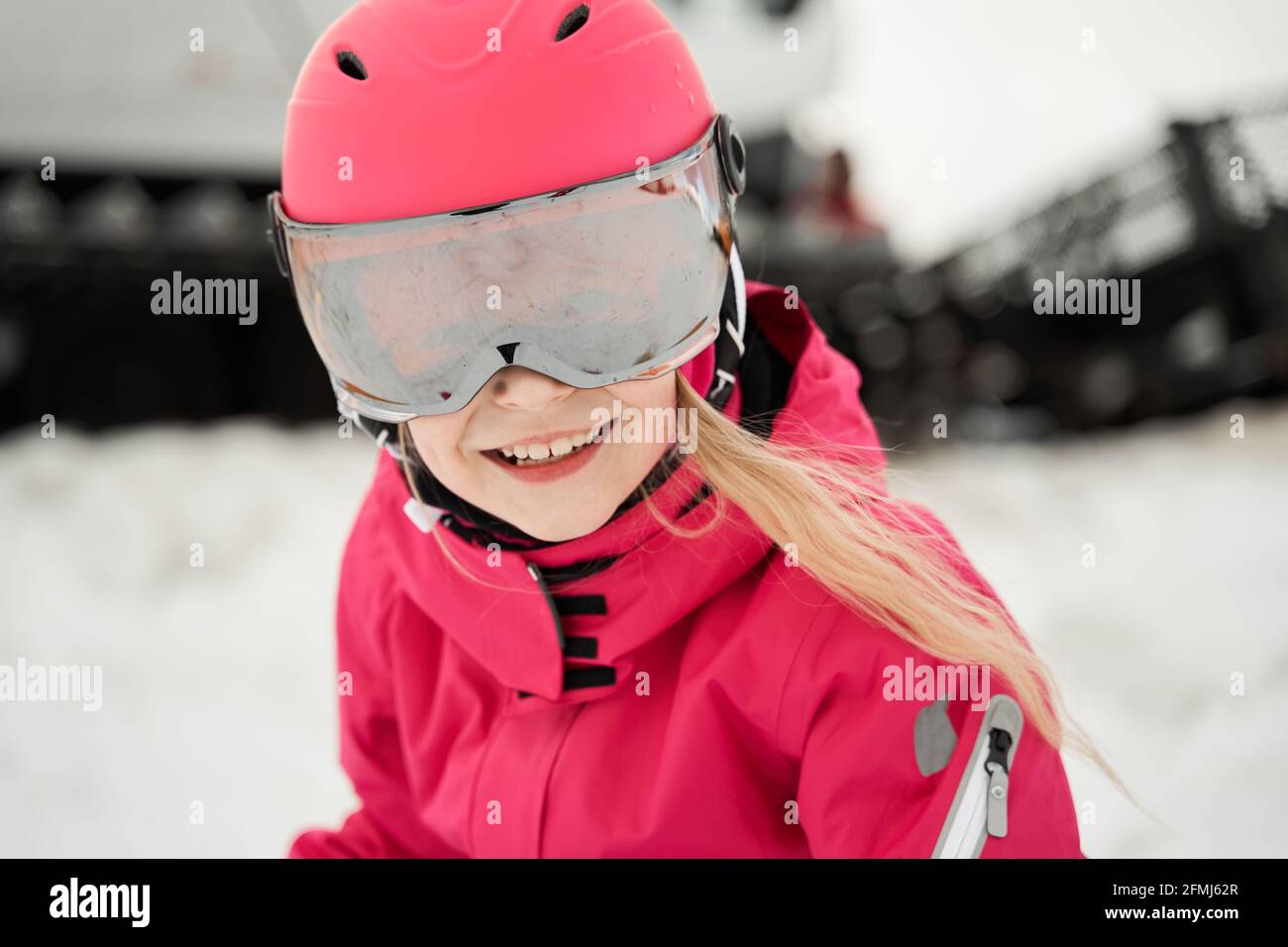 Positive cute girl in pink warm activewear goggles and helmet skiing ...