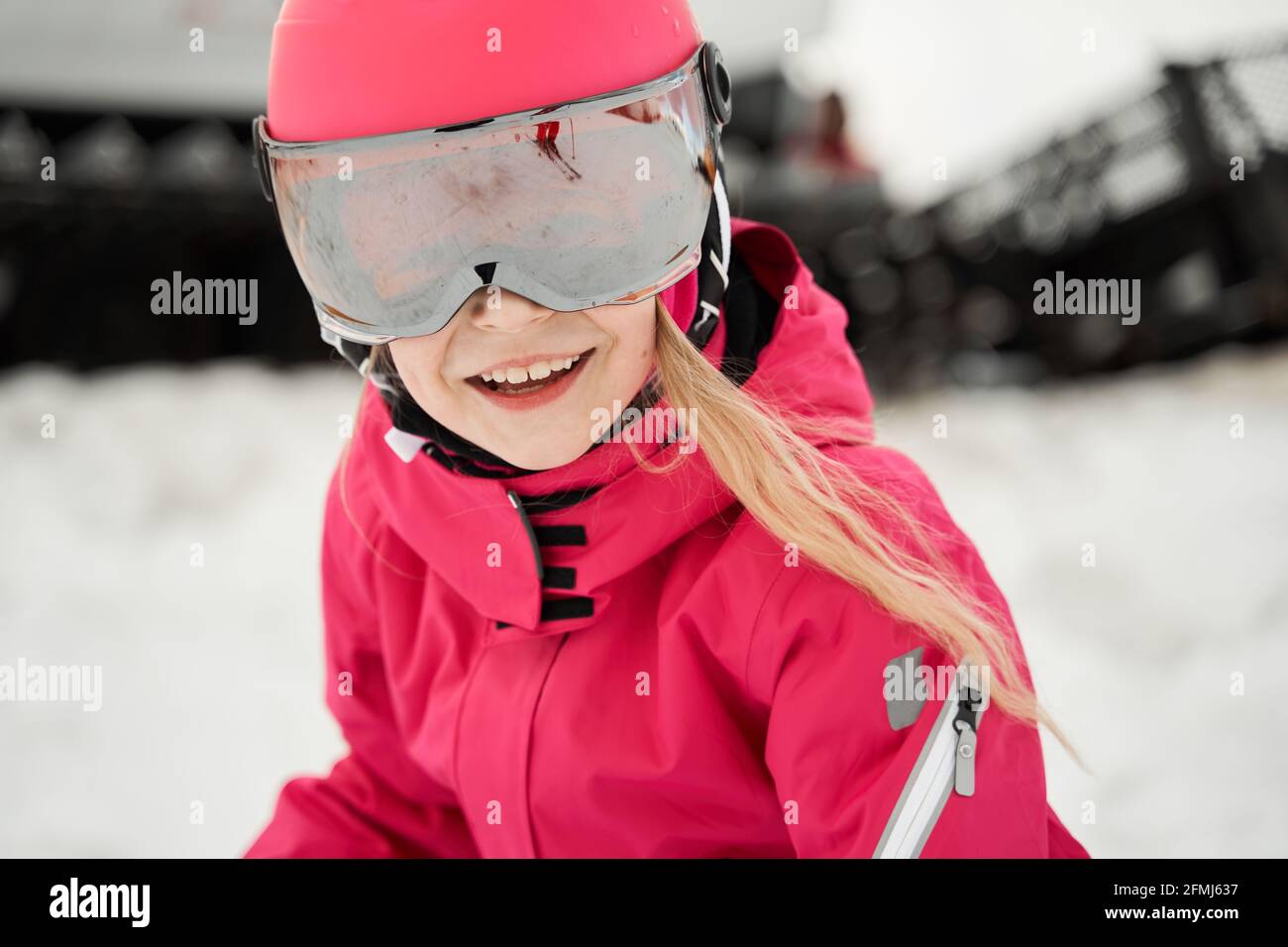 Positive cute girl in pink warm activewear goggles and helmet skiing ...