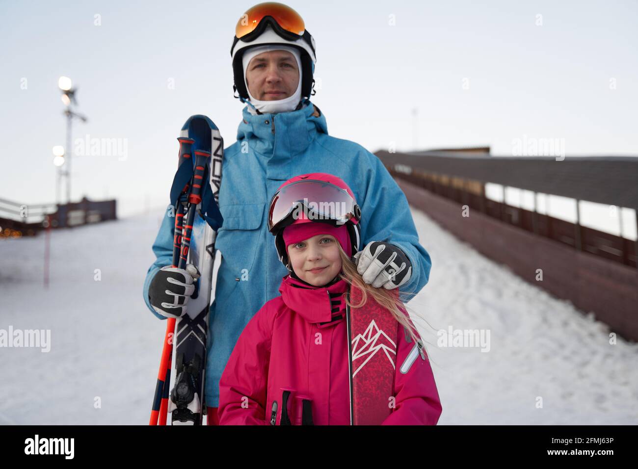 Glad father and daughter wearing warm sports clothes and helmets standing with skis on snowy hill slope and looking at camera contentedly Stock Photo