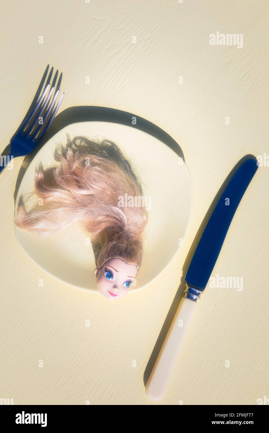 Head of blue eyed blonde haired doll on yellow plastic plate with knife and fork against yellow background. Concept of creepy, intrigue, mystery Stock Photo