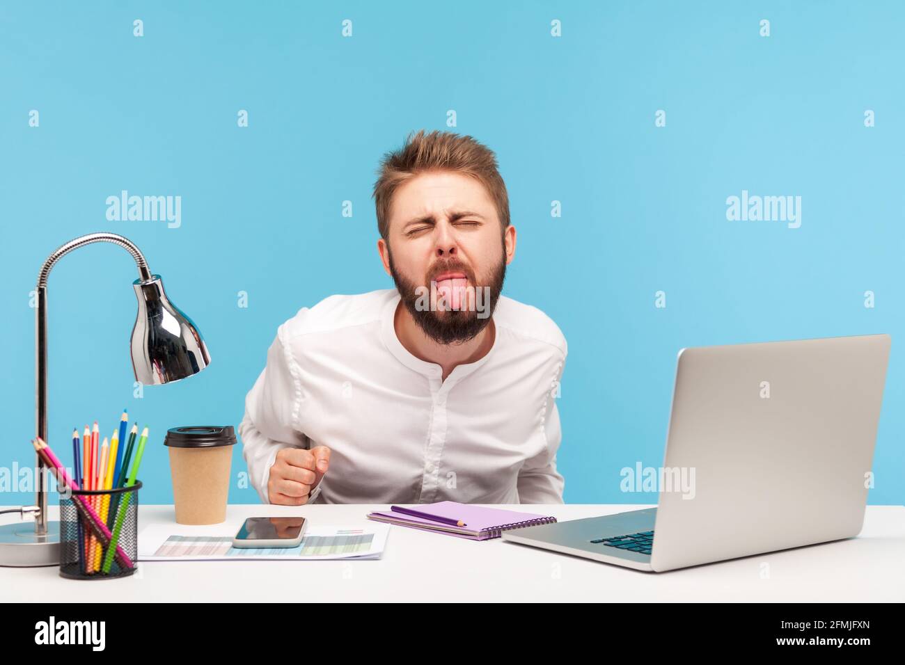 Crazy funny businessman sitting office workplace with laptop on desk, showing his tongue, looking at camera with naughty disobedient expression. Indoo Stock Photo