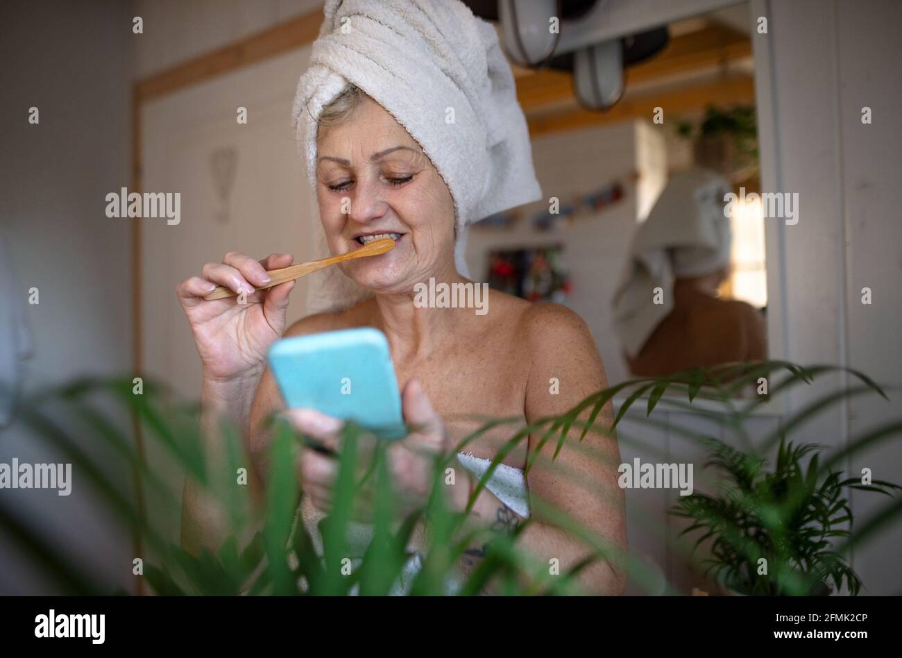 Senior woman with smartphone in bathroom brushing teeth at home, ecologic and sustainable lifestyle. Stock Photo
