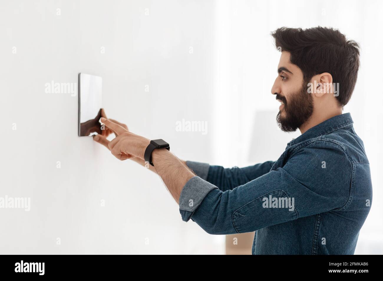 Bearded man controlling house with digital touch screen panel installed on light wall in the living room. Smart home and mobile application for managi Stock Photo