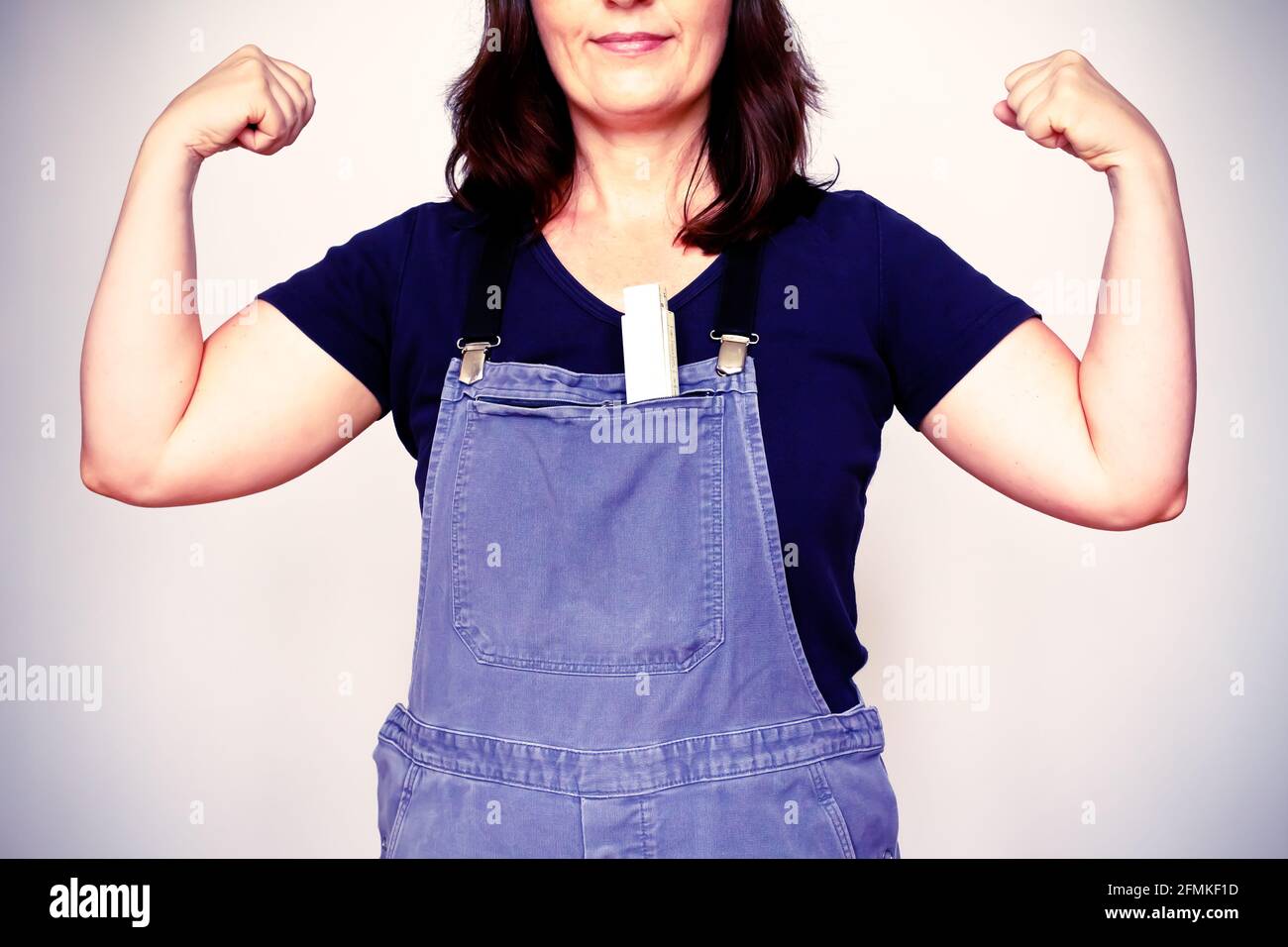 Self-confident woman in dungarees or bip trousers with a folding rule flexing her biceps muscles, filter effect. Stock Photo
