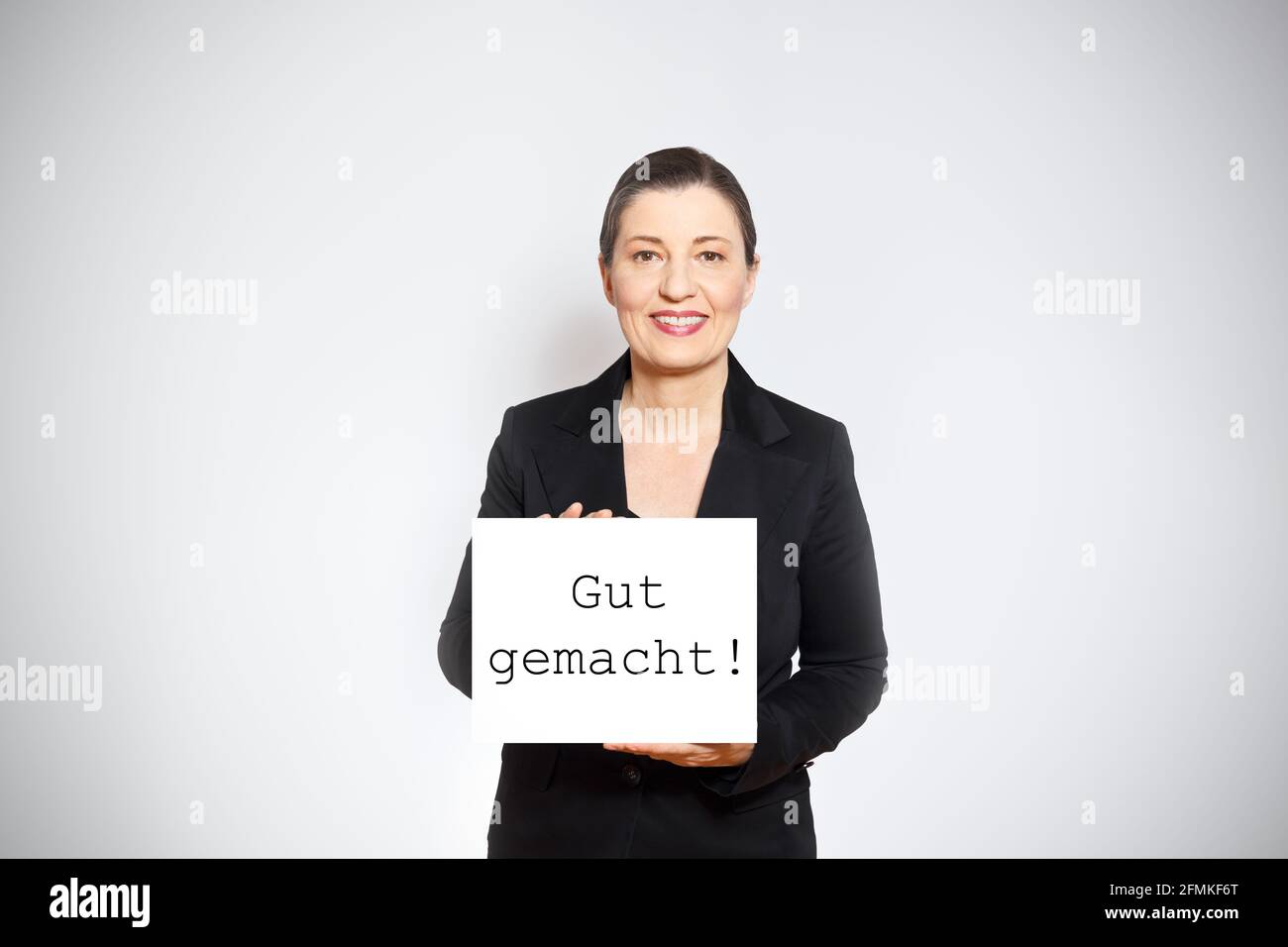 Smiling middle aged woman in black blazer holding up a sign with the text well done in german, white background. Stock Photo