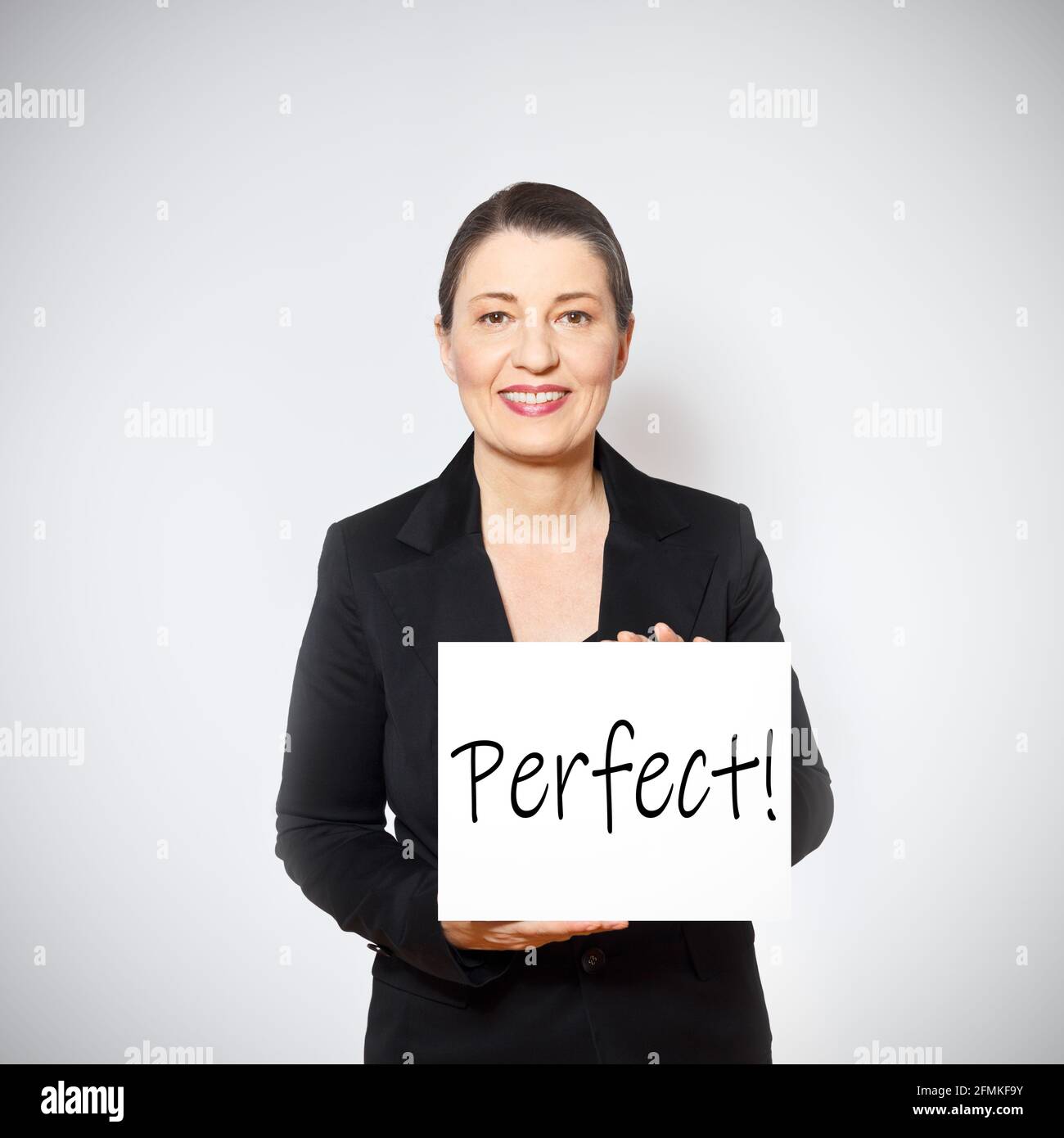 Smiling middle aged woman in black blazer holding up a sign with the praise: perfect, white background. Stock Photo