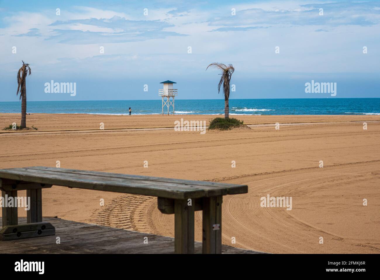 View of a wooden bench on a beach with dead palm trees and a lifeguard house in the background Stock Photo