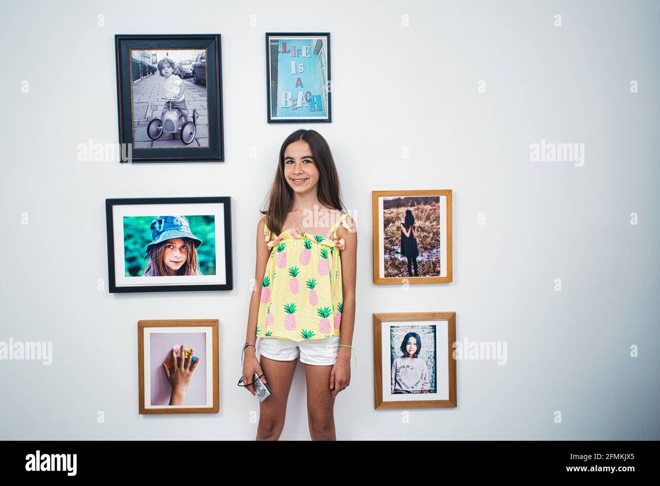young girl with camera standing against picture frames on wall at home. Stock Photo
