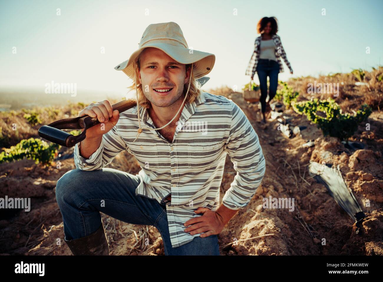 Handsome caucasian male farmer standing by crops holding shovel while female colleague works behind Stock Photo