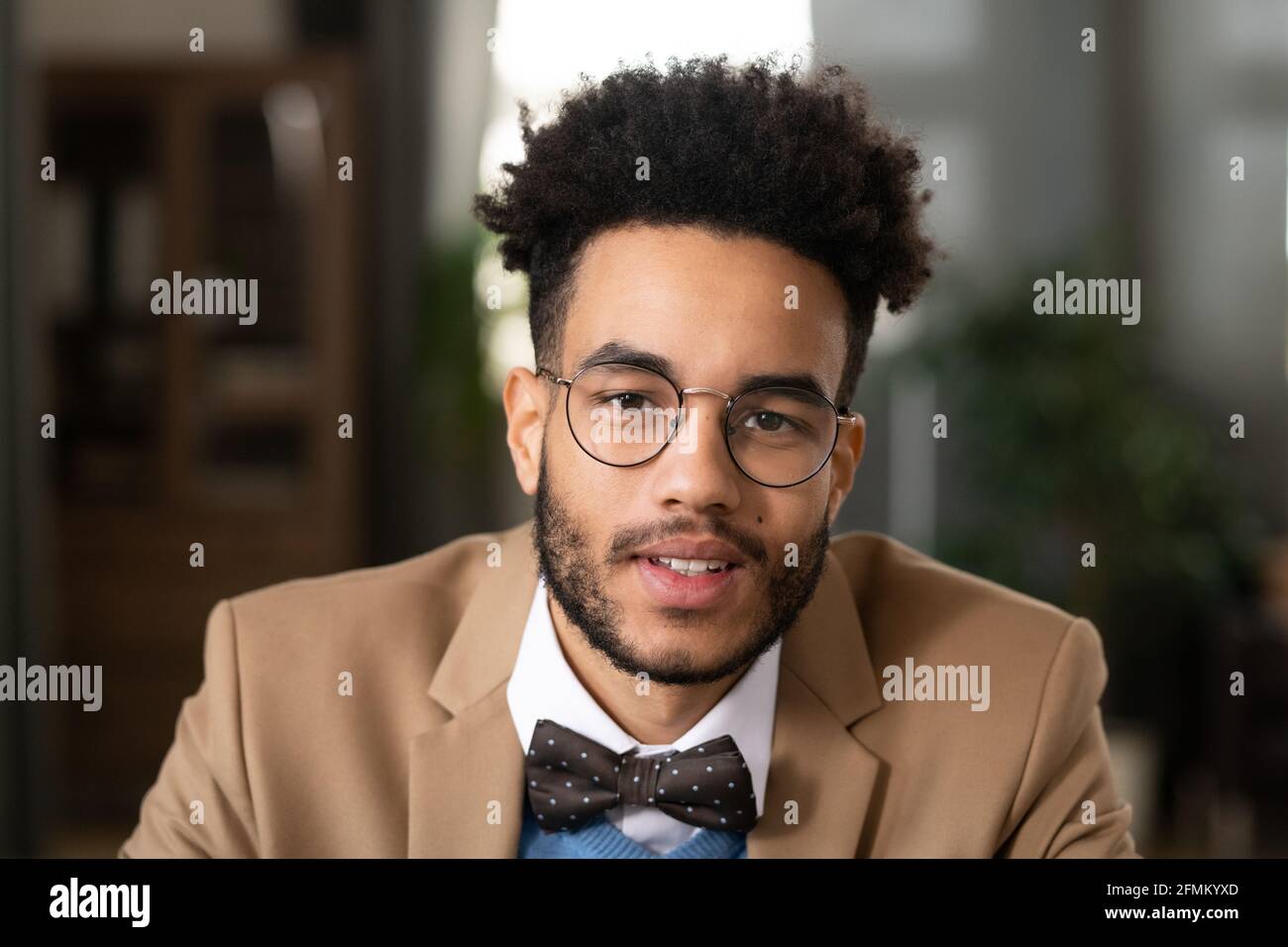 Portrait of positive handsome young black man with afro hairstyle wearing dotted bow tie and round-shaped glasses sitting in home office Stock Photo